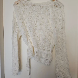 Vit stickad omlottkofta med knyt - Superfin vit stickad omlottkofta med knyt i midjan. Koftan har ett vackert spetsliknande mönster och långa ärmar. Perfekt för lager-på-lager och ger en romantisk vibe till din outfit.