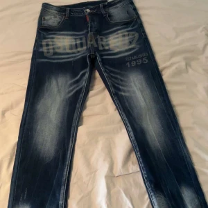 Dsquared2 jeans med tryck, blå  - Snygga blå jeans från Dsquared2 med stort tryck över framsidan och coola slitningar. Klassisk femficksmodell med knappgylf och tydliga kontrastsömmar. Modellen har en modern passform och är tillverkad i slitstarkt denimtyg.