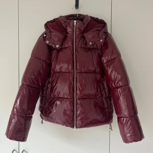 Vinröd pufferjacka med huva - Säljer min glansiga vinröda pufferjacka från Nelly, med huva och dragkedja framtill. Jackan har använts sparsamt och har inga tecken på användning. 