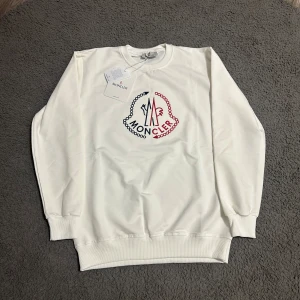 Vit sweatshirt från Moncler med broderi - Snygg vit sweatshirt från Moncler med stort broderat logomärke i rött och marinblått på bröstet. Tröjan har rund halsringning, ribbade muddar och är tillverkad i mjuk bomull. Perfekt för dig som gillar stilrena och exklusiva plagg.
