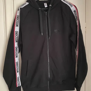 Svart hoodie från Moschino med logoband - Svart hoodie från Moschino med dragkedja och huva. Snygga vita logoband med svart Moschino-tryck och röda detaljer längs ärmarna. Långärmad modell med fickor framtill och ribbade muddar. Perfekt för en streetstyle-look. Nypris ca 3300kr.