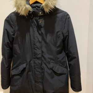 Svart parkasjacka med fuskpäls från Cross - Svart parkasjacka från Cross Sportswear med huva och fuskpälsdetalj. Jackan har långa ärmar, två stora fickor framtill och stängs med dragkedja och knappslå. Perfekt för kalla dagar med sin klassiska look och sköna passform.köpt för 1800kr på stadium 