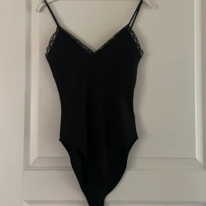 Svart body med spets urringning  - Snygg svart body med smala axelband och v-ringning. Toppen har fina spetsdetaljer längs urringningen och är figurnära med en enkel, stilren design. Perfekt att styla med jeans eller kjol för en trendig look.