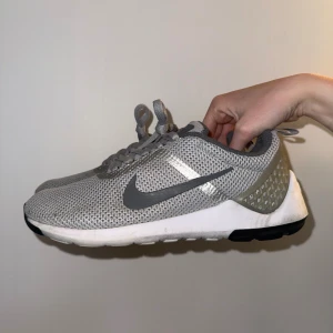 Gråa Nike skor - Snygga grå Nike sneakers med mesh-överdel och vita sulor. Skorna har mörkgrå swoosh på sidorna, snörning och detaljer i ljusgrått och silver. Baksidan har rutmönstrad plastdetalj och dragflik. Perfekta för dig som gillar sportig och clean stil.