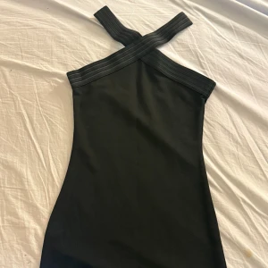 Svart kort klänning med korsade axelband - Snygg svart klänning med korsade axelband och figurnära passform. Klänningen är kort och har en stilren design utan mönster eller extra detaljer. Perfekt för dig som gillar minimalistisk stil och vill ha något som sticker ut med sin unika halsringning. Passar S-Xs