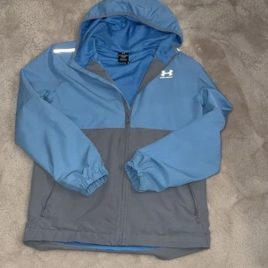 Blå och grå vindjacka Under Armour - Snygg vindjacka från Under Armour i blått och grått med huva och meshfoder. Jackan har dragkedja framtill, elastiska muddar och två sidofickor med dragkedja. Perfekt för aktiva dagar och sportiga outfits.