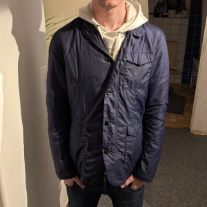 Mörkblå vindjacka med knappar - Säljer en mörkblå vindjacka med klassisk krage och flera fickor framtill. Jackan har knappar längs hela framsidan och är tillverkad i ett lätt, syntetiskt material som passar perfekt för blåsiga dagar. Snygg och enkel design med långärmade ärmar.