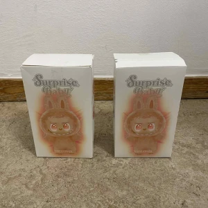 Surprise Baby Vinyl Plush Pendant Blind Box - Oöppnade Surprise Baby blind boxar med söta vinyl plush-figurer. Varje box innehåller en slumpmässig figur med olika teman som Happiness, Luck, Loyalty, Love, Serenity eller Hope. Perfekt som samlarobjekt eller present. Kartongerna är i gott skick med mindre slitage.