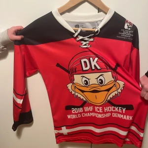Röd IIHF hockeytröja Danmark 2018 - Säljer en röd och svart officiell IIHF hockeytröja från VM 2018 i Danmark. Tröjan har coolt ankantryck med hjälm och texten DK, snörning i halsen och vita detaljer. Perfekt för hockeyfans som vill sticka ut på match eller i vardagen.