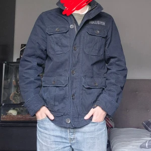 Mörkblå jacka från Hollister - Snygg mörkblå jacka från Hollister med fyra stora fickor framtill och tydlig logga på bröstet. Jackan har både dragkedja och knappar, upprätt krage och är tillverkad i bomull. Perfekt för dig som gillar en stilren och casual look.