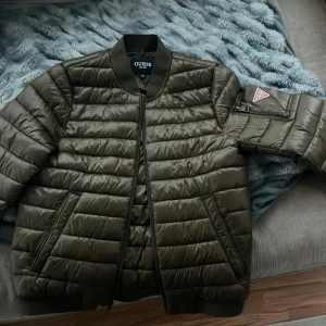 Olivgrön pufferjacka från Guess M - Säljer en olivgrön pufferjacka från Guess i storlek M. Jackan har quiltad design, dragkedja framtill, ribbade muddar och en ficka med Guess-logga på ärmen. Perfekt för kyliga dagar och riktigt snygg streetstil.