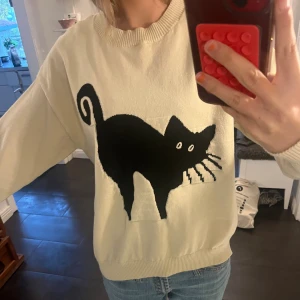 Beige stickad tröja med svart kattmotiv - Säljer en beige stickad tröja med ett stort svart kattmotiv på framsidan. Tröjan har rund halsringning, ribbade muddar och lång ärm. Perfekt för dig som gillar unika och lekfulla plagg med coola detaljer.🐈‍⬛