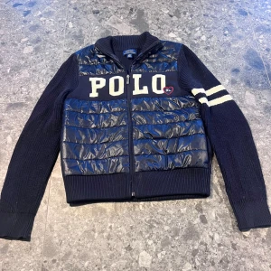 Blå stickad Polo Ralph Lauren-jacka - Mörkblå jacka från Polo Ralph Lauren med stickade ärmar och quiltad front i blankt material. Stora vita bokstäver 'POLO' framtill och två vita ränder på ena ärmen. Dragkedja hela vägen och ribbade muddar. Storlek M (8-10 år).