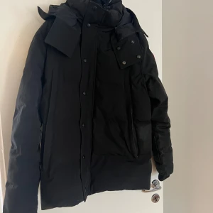 Svart dunjacka från Canada Goose - Säljer en svart dunjacka från Canada Goose med klassisk patch på ärmen. Jackan har hög krage, huva och flera fickor framtill. Perfekt för kalla dagar och har en clean, stilren look som funkar till allt. Stängs med både dragkedja och knappar. Storlek M päls finns om man vill ha med det pris kan diskuteras 