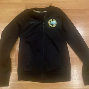 Svart Hammarby Craft ziptröja - Svart långärmad ziptröja från Craft med Hammarby IF-emblem på bröstet och stor Hammarby-tryck på ryggen. Tröjan har hel dragkedja framtill och är tillverkad i ett mjukt, sportigt material. Perfekt för dig som vill visa ditt stöd för Bajen.