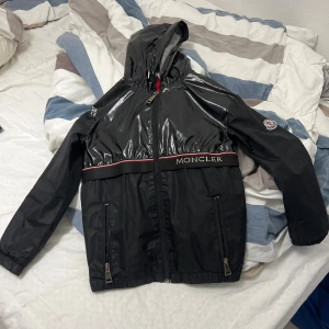 Svart vindjacka från Moncler med huva - Svart vindjacka från Moncler med glansig huva och överdel, dragkedja framtill och två fickor med dragkedja. Jackan har Moncler-logga på ärmen och ett broderat Moncler-band över bröstet. Insidan är grå med röd, vit och marinblå rand.