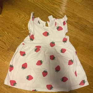 Vit klänning med jordgubbsmönster -74 cm  - Söt vit kortklänning med röda jordgubbar som mönster. Klänningen har ärmlös design med små volanger vid axlarna och rund hals. Tillverkad i mjuk bomull, perfekt för sommarens varma dagar. Passar dig som gillar lekfulla och färgglada plagg. #74 cm #9-12 månader #barnkläder #flicka 