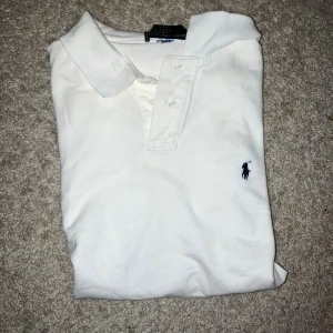 Vit polo från Ralph Lauren, storlek S - Vit klassisk polo från Ralph Lauren med broderad marinblå logga på bröstet. Skjortan har krage, knappar framtill och är gjord i mjuk bomull. Perfekt för en clean och stilren look.