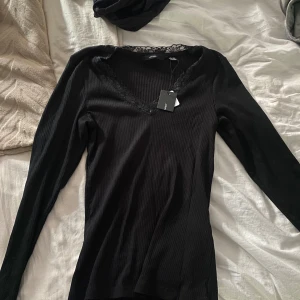 Svart ribbad topp med spets Vero Moda - Svart långärmad topp från Vero Moda i ribbad bomullsmix. Djup v-ringning med dekorativ spetskant runt halsen ger en snygg detalj. Perfekt att styla med jeans eller kjol för en trendig look.