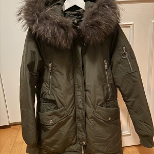 Rockandblue vinterkappa med pälskrage - Snygg olivgrön vinterjacka från Rockandblue med stor fluffig pälskant på huvan, dragkedjor på fickorna och ribbade muddar. Jackan har flera fickor och coola detaljer. Perfekt för kalla dagar. Pälskanten är äkta päls och är avtagbar.   