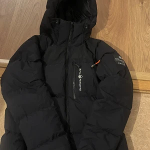 Svart dunjacka Sail Racing M - Säljer en svart dunjacka från Sail Racing i storlek medium. Jackan har huva, dragkedja framtill och en bröstficka med logga. Snygg och funktionell design med patch på ärmen och orange detalj vid dragkedjan. Perfekt för kalla dagar.