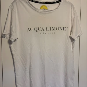 Vit Acqua Limone t-shirt med tryck - Vit t-shirt från Acqua Limone med klassiskt tryck framtill och stor gul citron på ryggen. Skön passform, rund hals och korta ärmar. Tillverkad i mjuk bomull, perfekt för en chill och avslappnad stil.