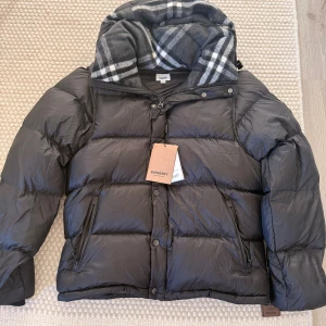Svart pufferjacka Burberry XL - Svart pufferjacka från Burberry i storlek XL. Jackan har en stor huva med grått och vitt rutigt mönster, dragkedja och knappar framtill samt två sidofickor med dragkedja. Materialet är polyester och polyamid, med detaljer i bomull och merinoull.