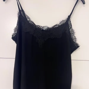 Svart linnetopp med spetsdetaljer - Snygg svart linnetopp med tunna axelband och v-ringning dekorerad med spets. Toppen har en elegant 