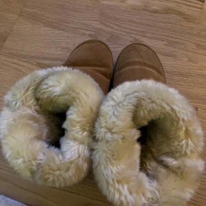 Bruna fårskinnsboots från Duffy - Mysiga bruna boots från Duffy med fluffigt ljust foder i fårskinnslook. Skorna har rund tå, platt sula och är tillverkade i mocka med en robust yttersula. Perfekta för kalla dagar när du vill hålla fötterna varma och sköna.