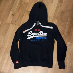 Mörkblå Superdry hoodie med tryck - Mörkblå hoodie från Superdry med stor vit, röd och blå logga på bröstet. Tröjan har huva med tjocka vita snören och känguruficka framtill. Mjuk insida och klassisk streetstil, perfekt för dig som gillar snygga och bekväma plagg. Riktigt mysig och stilren hoodie i väldigt bra skick då trycket är som nytt.   !OBS! väldigt liten i storlek! kommer troligtvis passa bra om du vanligtvis har S