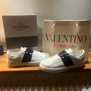 Valentino Garavani Open sneakers vit/svart - Säljer ett par Valentino Garavani Open sneakers i vitt skinn med bred svart rem över vristen och ikoniska nitar på hälen. Klassisk rund tå, platt sula och snygga detaljer. Perfekta för dig som vill ha en clean men lyxig look. Skolåda och bag följer med.