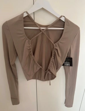 Beige croppad topp med snörning från Nelly - Säljer en beige croppad topp från Nelly med långa ärmar och snygg snörning baktill. Toppen har ett omlottliknande rynkat parti och tunna korsade band över ryggen. Materialet är mjukt och stretchigt, perfekt för att styla med jeans eller kjol.