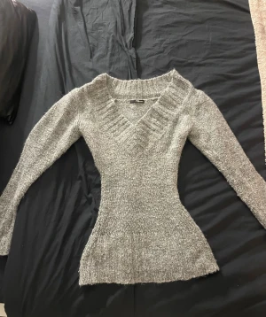 Grå stickad v-ringad tröja Jane Norman - Supersnygg grå stickad tröja från Jane Norman med v-ringning och lång ärm. Tröjan har en figurnära passform och bred ribbad kant vid hals, ärmslut och nederkant. Perfekt för kyliga dagar och enkel att styla med jeans eller kjol.