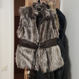 Brun pälsväst med bälte  - Säljer en fluffig grå pälsväst från Chic Rose med bred krage och snyggt bälte i midjan som knyts framtill. Västen har långa, mjuka pälsstrån i olika grå nyanser och detaljer i mörkbrunt syntetmaterial. Perfekt för att ge din outfit extra edge.