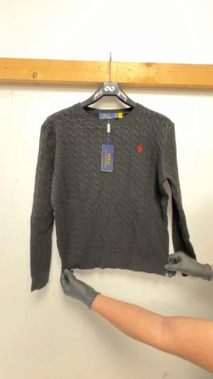 Svart stickad tröja - Snygg svart stickad tröja med klassiskt kabelstickat mönster och röd broderad logga på bröstet. Rund hals och långa ärmar, perfekt för lager-på-lager. Tröjan har en mjuk och skön känsla som passar till jeans eller chinos.