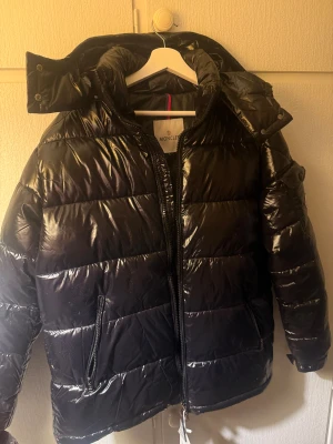 Moncler pufferjacka L - Svart pufferjacka från Moncler i storlek L med glansig finish och klassisk quiltad design. Jackan har huva med tryckknappar, dragkedja framtill och två sidofickor med dragkedja. Insidan har en cool Moncler-serieetikett. Perfekt för kalla dagar, avtagbar luva. Ej äkta!