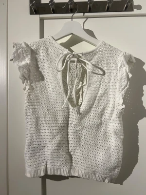 Vit spetsblus från Springfield - Supersöt vit blus från Springfield med broderade spetsdetaljer och volangärmar. Toppen har knytning i nacken och är ärmlös med vackert hålmönster och ribbad nederkant. 