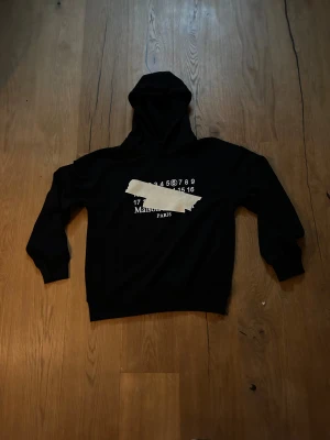 Maison Margiela svart hoodie med print - Svart hoodie från Maison Margiela med ikoniskt vitt print och tejpdetalj på bröstet. Tröjan har huva, ribbade muddar och lång ärm. Snyggt statement-plagg med cool Paris-vibe, perfekt för dig som gillar streetwear och designer. Strlk S men passar även M