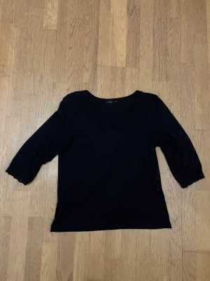 Svart blus med spets från Fransa - Svart blus från Fransa med v-ringning och fina spetsdetaljer längs halsringningen och ärmsluten. Modellen har trekvartsärm och är i mjukt, lätt material som känns skönt mot huden. Perfekt för dig som gillar stilrena och enkla plagg med en twist.