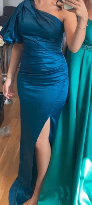 Blå långklänning med slits - Snygg blå långklänning i satin med oneshoulder och puffärm. Klänningen har en hög slits på sidan som ger en cool och elegant vibe. Perfekt för bal eller fest, och den glansiga ytan gör att du sticker ut. Passformen är slim och tyget sitter snyggt mot kroppen. Endast använd en gång , passar storlek 34/36 