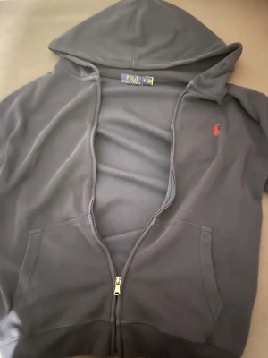 Blå hoodie från Polo Ralph Lauren - Säljer en klassisk marinblå hoodie från Polo Ralph Lauren med röd broderad logga på bröstet. Tröjan har huva, dragkedja framtill och två fickor. Perfekt för chill dagar och snygg till jeans eller joggers.