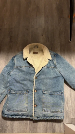 Blå teddyfodrad jeansjacka från ASOS XL - Säljer en blå jeansjacka från ASOS i storlek XL med mjukt teddyfoder i krage och insida. Jackan har stora fickor framtill, knäppning med knappar och en avslappnad passform. Perfekt för kyliga dagar när du vill ha både stil och komfort.