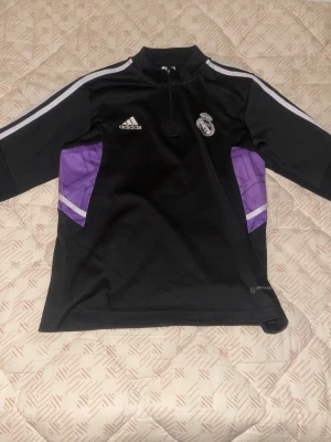 Real Madrid träningströja Adidas - Säljer en svart långärmad Real Madrid träningströja från Adidas med dragkedja vid halsen. Tröjan har vita ränder på ärmarna och lila detaljer på sidorna. Perfekt för fotbollsträning eller som supporterplagg.