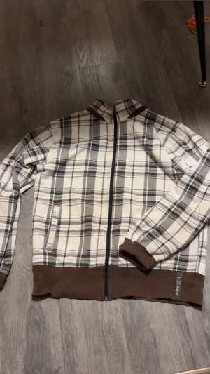 Rutig vindjacka från Black Temple - Snygg rutig vindjacka från Black Temple i vitt, grått och brunt. Jackan har hel dragkedja, bruna muddar och nederkant samt två fickor framtill. Perfekt för dig som gillar streetstyle och vill sticka ut med ett klassiskt rutmönster.