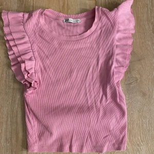 Rosa volangtopp från Zara - Supersöt rosa topp från Zara med ribbad struktur och volangdetaljer längs axlarna. Toppen har rund halsringning och är ärmlös, perfekt för en fräsch och trendig look.