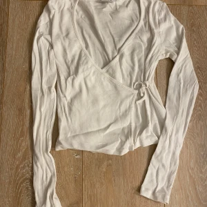 Vit omlottopp med knyt och v-ringning - Säljer en vit omlottopp med lång ärm och v-ringning. Toppen har ribbad struktur och knyt i sidan för en snygg detalj. Perfekt för dig som gillar en enkel men trendig stil.