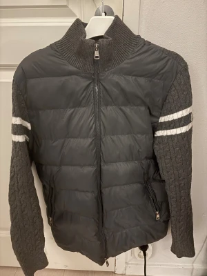 Grå Moncler pufferjacka med stickade ärmar - Säljer en grå pufferjacka från Moncler med stickade, kabelmönstrade ärmar och ribbad krage. Jackan har vita ränder på ärmarna, två dragkedjefickor och Moncler-logga på vänster ärm. Perfekt för dig som vill ha en unik och snygg jacka med coola detaljer.