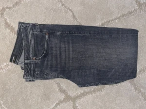 Svarta jeans Tapered Mike Jack & Jones - Snygga svarta jeans från Jack & Jones, modell Tapered Mike. Jeansen har klassisk femficksdesign, knappgylf och coola slitningar för en avslappnad vibe. Passformen är tapered med smalare ben och de är tillverkade i mjukt jeanstyg för skön komfort.. regular fit i storlek 31W/32L. Helt nytt skick