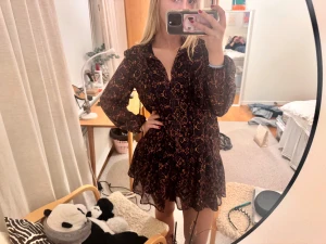 Mörk mönstrad kortklänning med volang - Supersöt kortklänning med färgglatt mönster i orange och lila. Klänningen har långa, ärmar, v-ringning och volangkant nertill. Det är luftigt material som känns lätt och lite flowy. Perfekt till jul eller nyår!❤️❤️