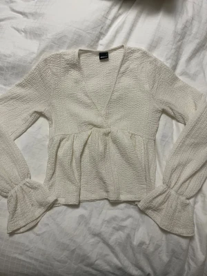 Vit blus med volang från Gina Tricot - Supersöt vit blus från Gina Tricot i storlek S. Blusen har en v-ringad hals, långärmade ärmar med volangdetalj vid mudden och ett strukturerat tyg. Passformen är normal!🤍🤍🤍🤍Perfekt till ett par jeans😍
